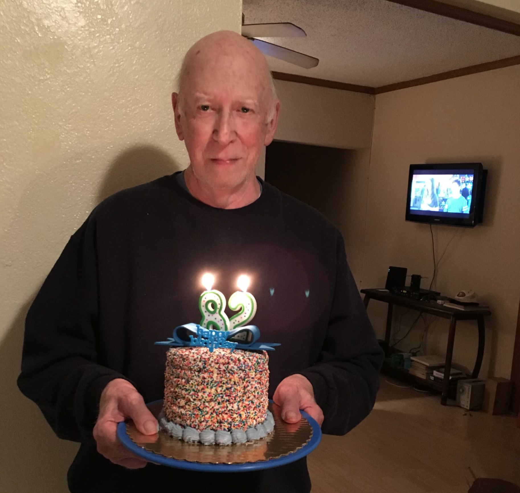 A balding white man holding a birthday cake which says "82" on it.